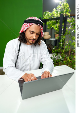 Young Arab man in traditional dress thobe working on computer in office Young Arab man in traditional dress thobe working on computer in office 109768189