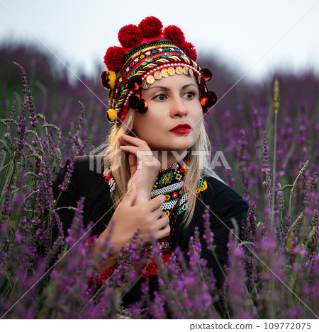 A girl in a chelsea headdress and a black dress decorated with red embroidery sits between lavender bushes. A girl in a chelsea headdress and a black dress decorated with red embroidery sits between lavender bushes. 109772075