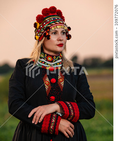 A girl in a chelsea headdress and a black dress decorated with red embroidery on the background of a field and sky. 109772076