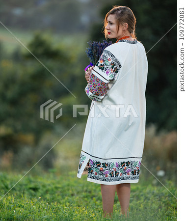 A young girl wearing a long embroidered shirt holds a bouquet of lavender in her hands. 109772081