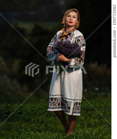 A young girl wearing a long embroidered shirt holds a bouquet of lavender in her hands. A young girl wearing a long embroidered shirt holds a bouquet of lavender in her hands. 109772082