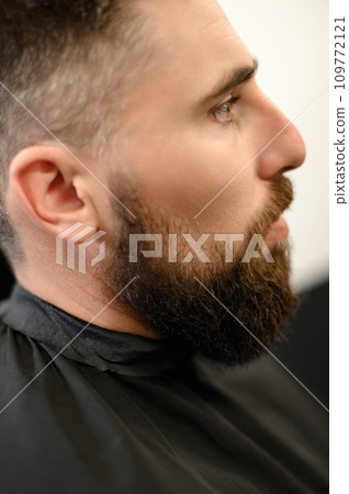 A young man with a beard in a black cape sits on a chair in a barbershop. The client is waiting for a haircut of the beard. 109772121