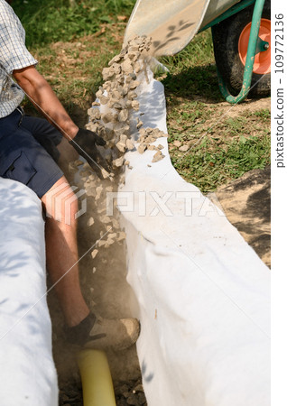 Man pours rubble from a wheelbarrow into a trench. Drainage works for drainage of ground water around the field. Man pours rubble from a wheelbarrow into a trench. Drainage works for drainage of ground water around the field. 109772136