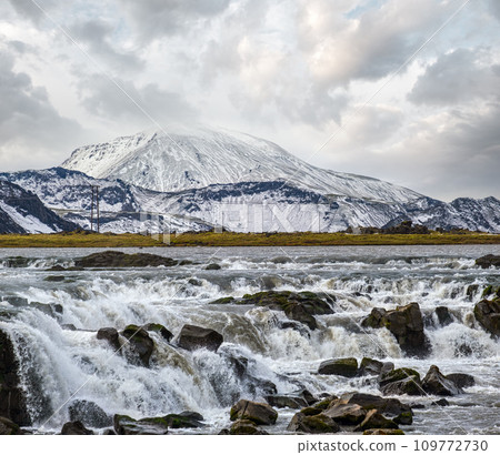 Season changing in southern Highlands of Iceland. Picturesque waterfal Tungnaarfellsfoss panoramic autumn view.  Landmannalaugar mountains under snow cover in far. 109772730