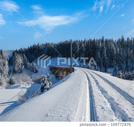 Stone viaduct (arch bridge) on railway through mountain snowy fir forest. Snow drifts on wayside and hoarfrost on trees and electric line wires. Stone viaduct (arch bridge) on railway through mountain snowy fir forest. Snow drifts on wayside and hoarfrost on trees and electric line wires. 109772976