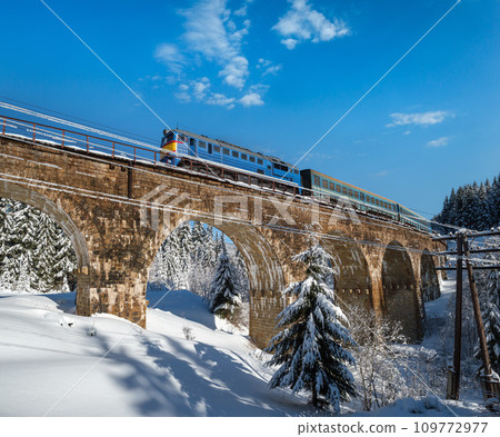 Stone viaduct (arch bridge) on railway through mountain snowy fir forest and locomotive with a passenger train. Snow drifts on wayside and hoarfrost on trees and electric line wires. Stone viaduct (arch bridge) on railway through mountain snowy fir forest and locomotive with a passenger train. Snow drifts on wayside and hoarfrost on trees and electric line wires. 109772977