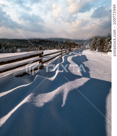 Picturesque waved shadows on snow from wood fence. Alpine mountain winter hamlet outskirts, snowy path, fir forest. High resolution image with great depth of field. 109772994