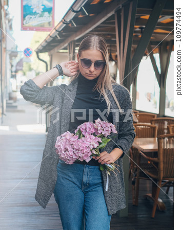 Beautiful caucasian girl with blond hair in a gray coat and blue jeans with a bouquet of pink hydrangeas and with the sunglasses with a city view Beautiful caucasian girl with blond hair in a gray coat and blue jeans with a bouquet of pink hydrangeas and with the sunglasses with a city view 109774494