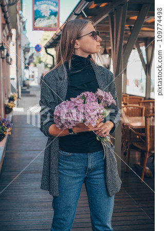 Beautiful caucasian girl with blond hair in a gray coat and blue jeans with a bouquet of pink hydrangeas and with the sunglasses with a city view Beautiful caucasian girl with blond hair in a gray coat and blue jeans with a bouquet of pink hydrangeas and with the sunglasses with a city view 109774498