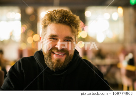 Bearded guy programmer or an entrepreneur in glasses in a stylish cafe looks at the camera and smiles. Freelancer works remotely. Online communication video call and small business. Bearded guy programmer or an entrepreneur in glasses in a stylish cafe looks at the camera and smiles. Freelancer works remotely. Online communication video call and small business. 109774963