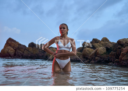 Attractive woman enjoys nighttime swim in ocean under crimson illumination. Fashionable lady in swimwear poses during an evening ocean dip. Attractive woman enjoys nighttime swim in ocean under crimson illumination. Fashionable lady in swimwear poses during an evening ocean dip. 109775254
