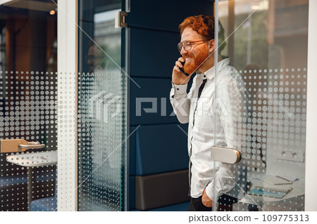 Bearded male freelancer in eyeglasses is talking phone standing in modern coworking space Bearded male freelancer in eyeglasses is talking phone standing in modern coworking space 109775313