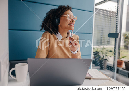 Smiling female manager in headphones listen music and looks away while working laptop in office Smiling female manager in headphones listen music and looks away while working laptop in office 109775431
