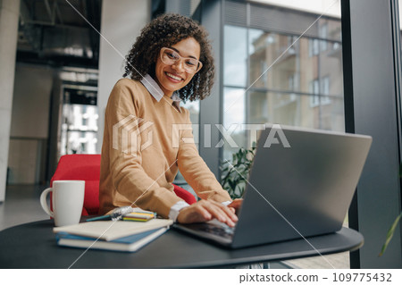 Pretty female manager in eyeglasses working on laptop while sitting in modern office near window 109775432
