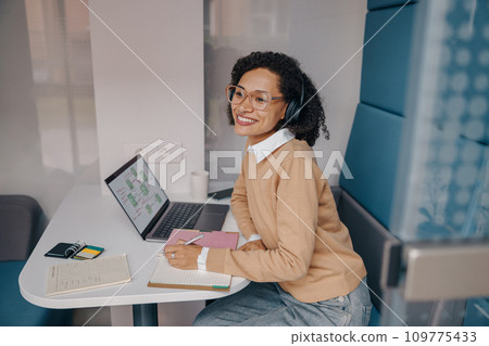 Smiling female analyst in eyeglasses working on laptop and making notes while sitting in cozy office Smiling female analyst in eyeglasses working on laptop and making notes while sitting in cozy office 109775433