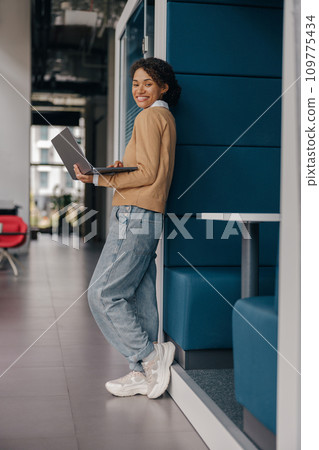 Stylish female manager using laptop while standing on office background and looks camera Stylish female manager using laptop while standing on office background and looks camera 109775434
