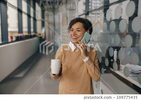 Smiling female manager with coffee cup is talking phone while standing on modern office background Smiling female manager with coffee cup is talking phone while standing on modern office background 109775435