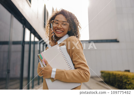 Pretty female entrepreneur is talking phone while standing with laptop on modern building background Pretty female entrepreneur is talking phone while standing with laptop on modern building background 109775482