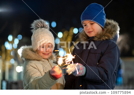 Two cute young children, boy and girl in warm winter clothing holding burning sparkler fireworks on dark night outdoors bokeh background. New Year and Christmas celebration concept. 109775894