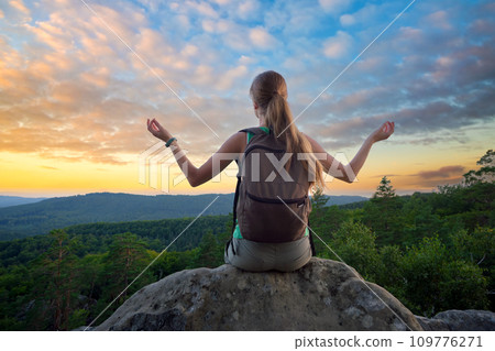 Sportive female hiker sitting alone taking a meditation break on hillside trail. Lonely woman enjoying view of evening nature from rocky cliff on wilderness path. Active lifestyle concept 109776271