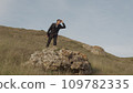 A man in a classic suit stands on a rock protruding from a hill and folds his hands with his glasses and peers into the distance 109782335