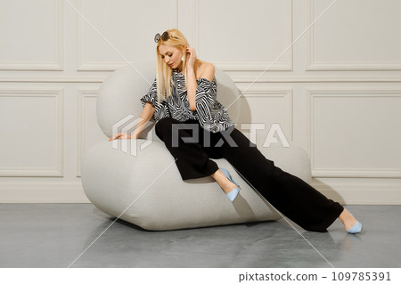 A beautiful blond-haired woman in fashionable boho style clothes sits with her legs crossed in a soft bean bag chair A beautiful blond-haired woman in fashionable boho style clothes sits with her legs crossed in a soft bean bag chair 109785391