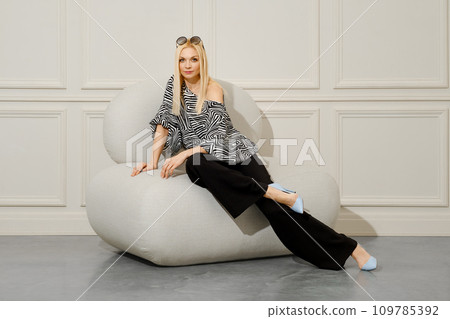 A fashionable woman sits in a soft chair, wearing wide trousers and a patterned blouse 109785392