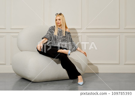 A beautiful, stylish woman in boho blouse and loose trousers sits comfortably in a soft chair. 109785393