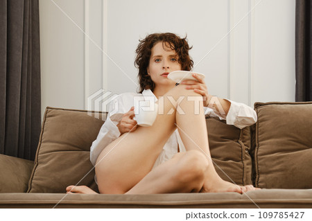dreamy lonely young woman sitting on sofa with cup of cappuccino on her knee 109785427
