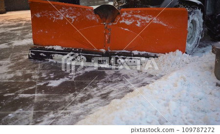 A bucket close-up cleans the sidewalk from snow. The tractor cleans the pavement in winter. Tractor cleaning the road from the snow. Excavator cleans the streets of large amounts of snow in city. 109787272