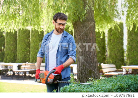 Caucasian man in protective glasses cutting boxwood bush with hedge trimmer outside. Front view of professional gardener using modern equipment, while taking care of plants. Concept of seasonal work. Caucasian man in protective glasses cutting boxwood bush with hedge trimmer outside. Front view of professional gardener using modern equipment, while taking care of plants. Concept of seasonal work. 109787681