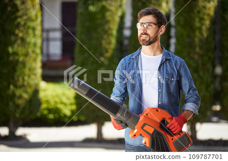Attractive bearded male worker carrying modern cordless leaf blower on backyard. Portrait of smiling gardener in glasses operating powerful equipment on blurred background. Concept of seasonal work. 109787701