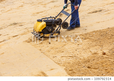 Worker using vibratory plate compactor for compaction sand during path construction. 109792314