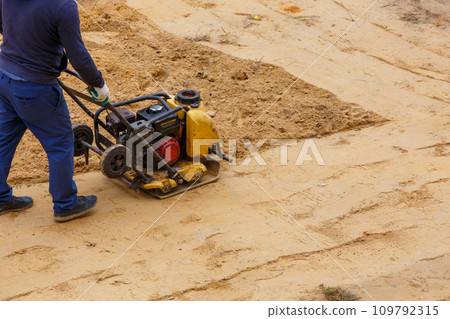 Worker using vibratory plate compactor for compaction sand during path construction. Worker using vibratory plate compactor for compaction sand during path construction. 109792315