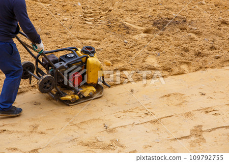 Worker using vibratory plate compactor for compaction sand during path construction. Worker using vibratory plate compactor for compaction sand during path construction. 109792755