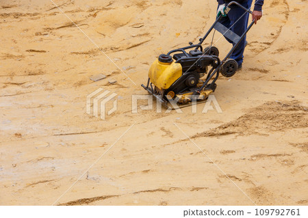 Worker using vibratory plate compactor for compaction sand during path construction. Worker using vibratory plate compactor for compaction sand during path construction. 109792761