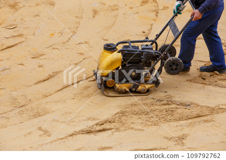 Worker using vibratory plate compactor for compaction sand during path construction. Worker using vibratory plate compactor for compaction sand during path construction. 109792762
