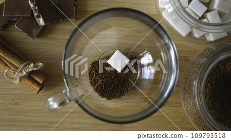Close up top view of a woman hand putting granulated instant coffee and sugar into the mug. Concept. Making transparent cup of coffee placed on brown wooden surface with chocolate and cinnamon. 109793518
