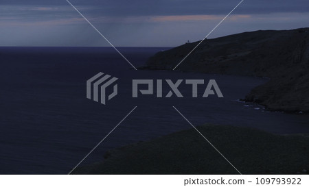 Aerial view of the long grey cliff covered with shrubs and the lighthouse near sea against blue with pink evening sky. Shot. Beautiful evening marine scenery 109793922