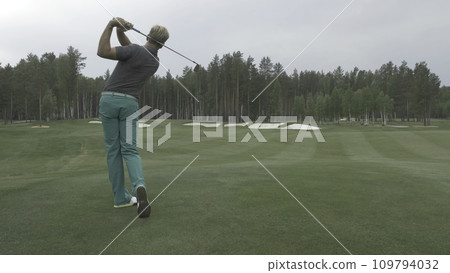 Golfer Hitting Ball with Club on Beautiful Golf Course. Golfer hits an fairway shot towards the club house. Man hitting driver on a golf course in the sun. Professional male golf player Golfer Hitting Ball with Club on Beautiful Golf Course. Golfer hits an fairway shot towards the club house. Man hitting driver on a golf course in the sun. Professional male golf player 109794032