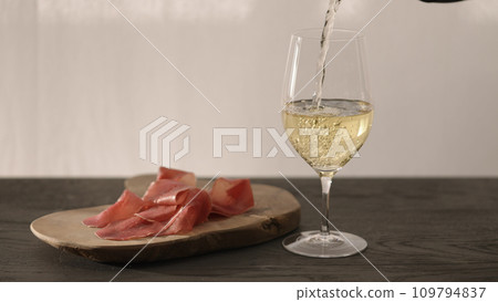 pour white whine in wine glass on oak table with bresaola on olive board 109794837