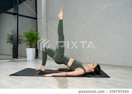 Flexible sporty woman wearing sage color gym clothes resting in One Legged Bridge pose on yoga mat. Side view of focused female with closed eyes practicing yoga at home. Concept of yoga practice. 109795840