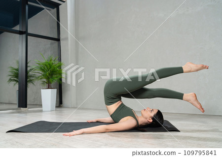 Strong flexible woman wearing sportswear practicing supine yoga pose at home. Side view of focused caucasian female trying to do halasana on yoga mat. Concept of yoga practice, lifestyle. Strong flexible woman wearing sportswear practicing supine yoga pose at home. Side view of focused caucasian female trying to do halasana on yoga mat. Concept of yoga practice, lifestyle. 109795841