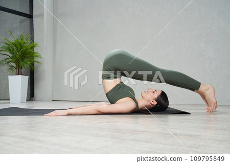Calm flexible woman wearing olive gym clothes in asana halasana on yoga mat. Side view of smiling young female with closed eyes practicing yoga at home. Concept of yoga practice, lifestyle. Calm flexible woman wearing olive gym clothes in asana halasana on yoga mat. Side view of smiling young female with closed eyes practicing yoga at home. Concept of yoga practice, lifestyle. 109795849