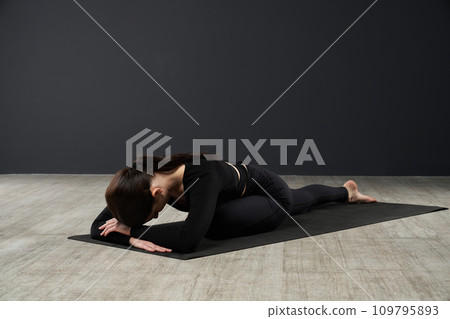Unrecognizable slim woman sitting in Pigeon pose of yoga folding forward on mat. Side view of young female practicing Eka Pada Rajakapotasana during yoga class in studio. Yoga lifestyle concept. Unrecognizable slim woman sitting in Pigeon pose of yoga folding forward on mat. Side view of young female practicing Eka Pada Rajakapotasana during yoga class in studio. Yoga lifestyle concept. 109795893