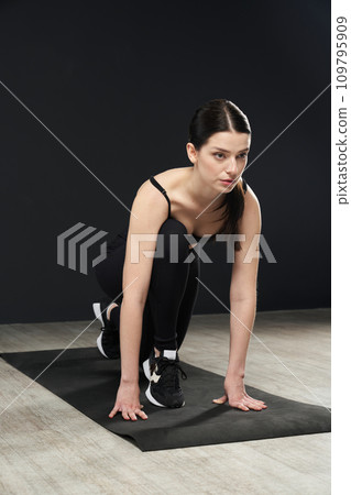 Athletic focused woman in black sportswear standing in crouch start pose on rubber yoga mat. Close up view of beautiful brunette sportswoman exercising in studio. Concept of sport, training. 109795909