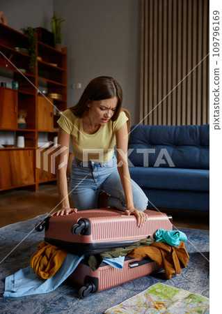 Young woman struggling with overload travel suitcase while preparing for trip 109796169