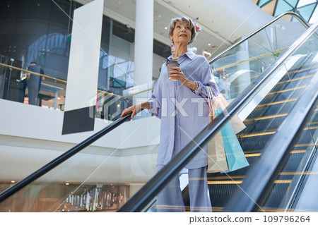 Beautiful aged woman in casual clothes with takeaway coffee on escalator Beautiful aged woman in casual clothes with takeaway coffee on escalator 109796264