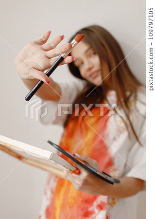 Young female artist posing holding brushes and painting in hands. Woman wearing white and red t-shirt. Brunette girl standing in a room with white wall. 109797105