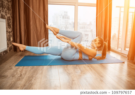 Young athletic attractive woman practicing yoga doing exercises. Works out at home or in a yoga studio, sportswear, blue pants and a full-length top indoors. Healthy lifestyle concept Young athletic attractive woman practicing yoga doing exercises. Works out at home or in a yoga studio, sportswear, blue pants and a full-length top indoors. Healthy lifestyle concept 109797657